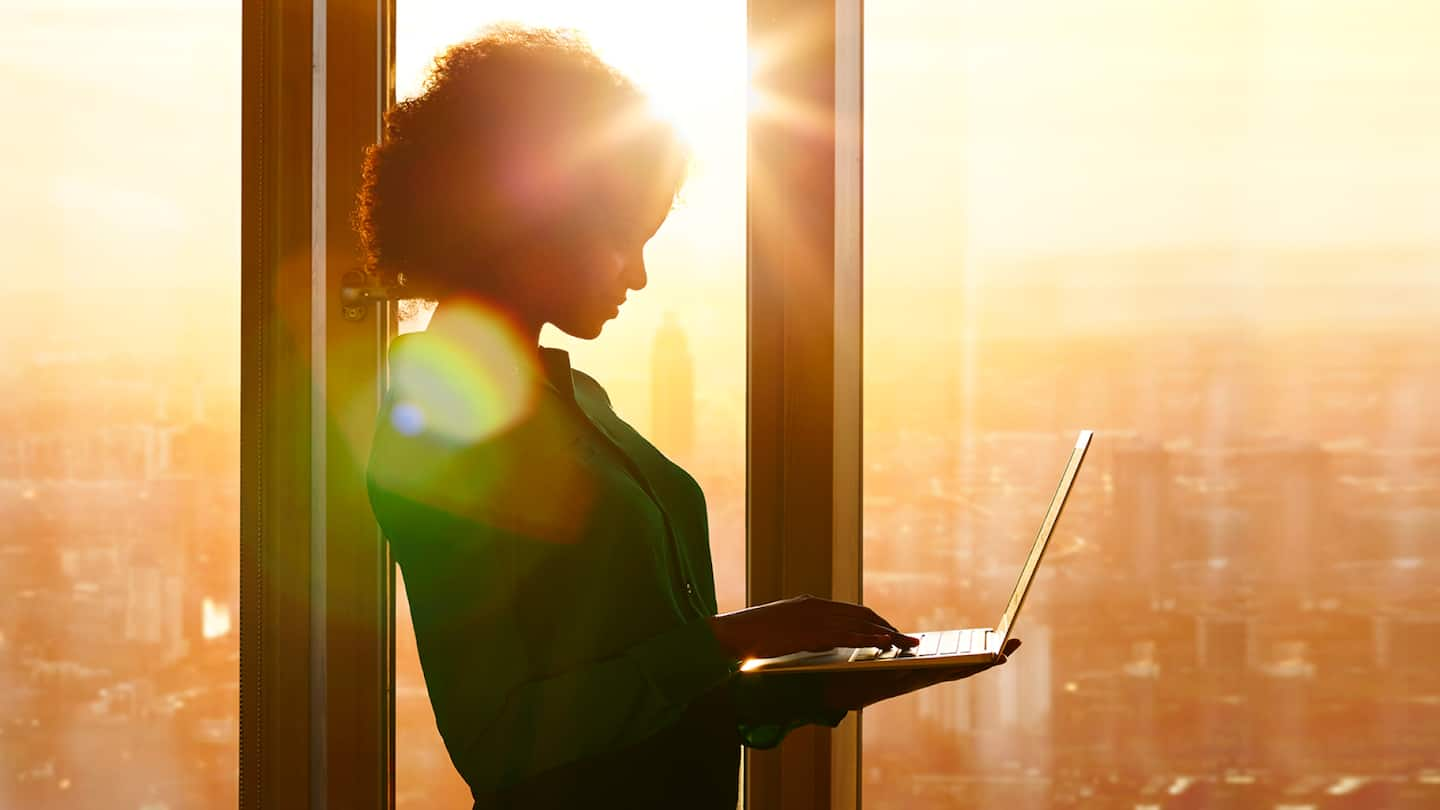 Silhouette of a woman at twilight standing in front of a skyscraper window using cloud computing services on her laptop