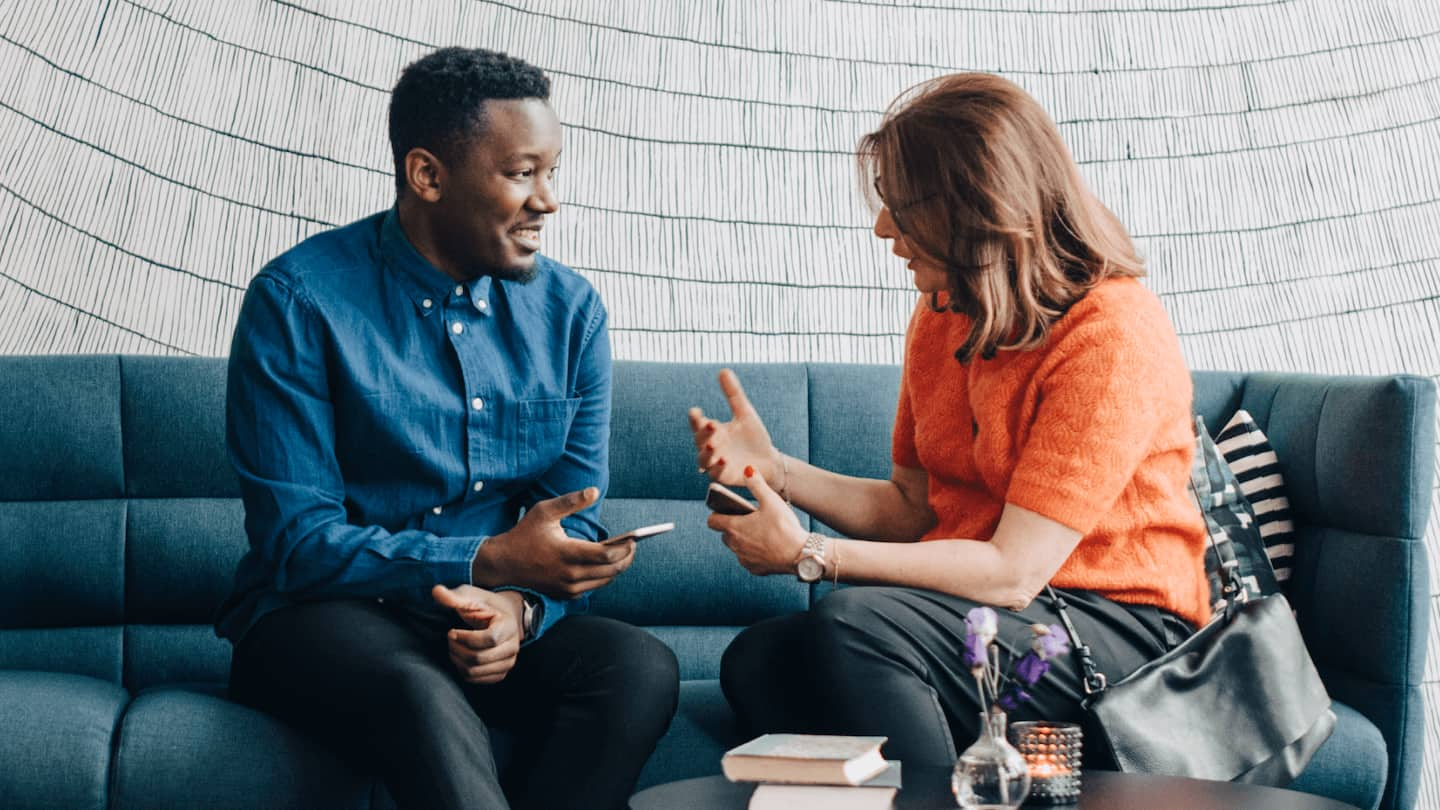 A male wearing a blue shirt and a female wearing an orange shirt sit next to each other on a couch having a discussion. 